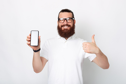 This is a good offer. Check it out. Bearded man is holding phone and showing thumb up over white background.