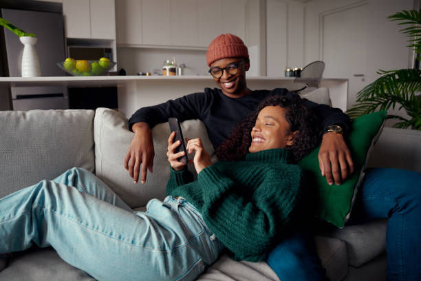 istock Multi-ethnic couple laughing, looking at text on phone while on the couch in modern apartment 1348046356