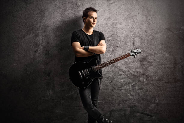 Young man with an electric guitar on a strap leaning on a gray rugged wall