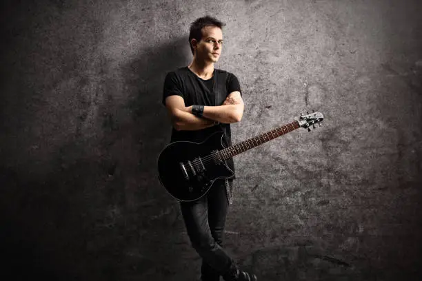 Photo of Young man with an electric guitar on a strap leaning on a rugged wall