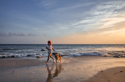 Happy red-haired woman in casual summer style running on the beach with her dog