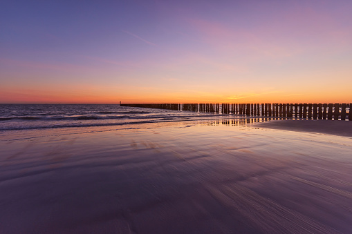 A row of poles going into the water on the beach of Zeeland, the Netherlands. The sun is setting and creates a beautifully colored sky.  The water reflects its surroundings.