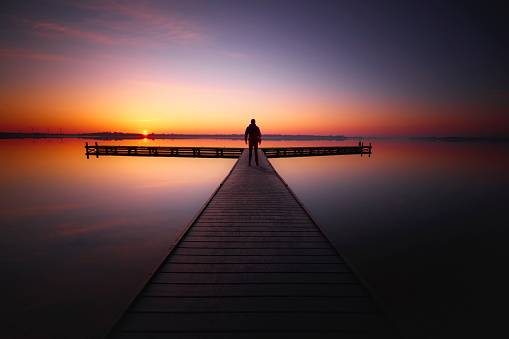 Someone is walking on a wooden jetty over the water during sunset. The sky is beautifully colored and the water reflects it because it is very quiet. On the other side of the water is land.