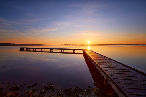 There is a wooden jetty over the water during sunset. The sky is beautifully colored and there is no one to be seen. The water is very quiet and reflects the sky and on the other side of the water is land.