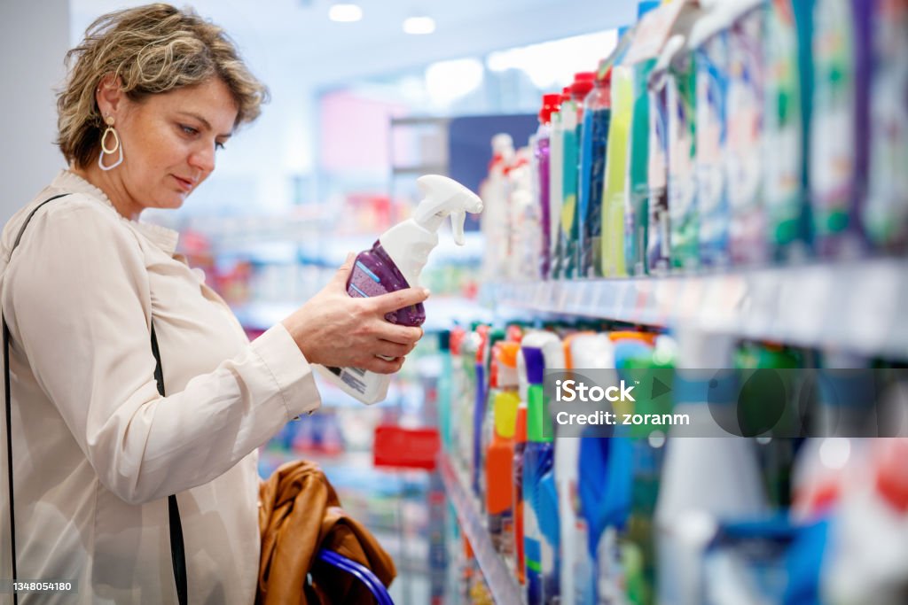 Woman choosing domestic cleaning product by the supermarket shelf Woman standing by the supermarket shelf with household hygiene products, holding cleaning agent and reading label Cleaning Product Stock Photo