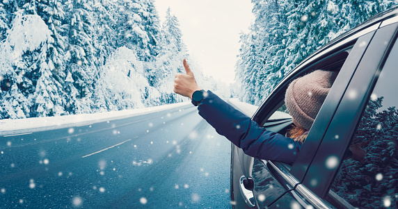 Happy woman in the car gesture finger up on the snowy background. Auto on the winter country road in the forest. Concept of the family vacation and tourism.