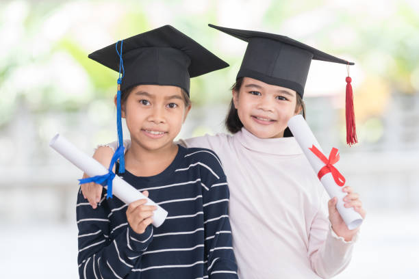 Two happy Asian female school kid graduates with a graduation cap holds a rolled certificate stock photo
