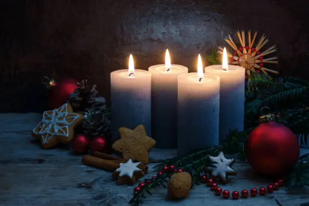 Four Advent candles with Christmas decoration, baubles and cookies on rustic wooden planks against a dark blue background with copy space, selected focus, narrow depth of field