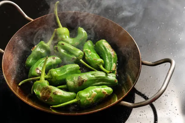 Steaming peppers de padron or green pimientos in a hot frying pan on the stove for a Spanish tapa or appetizer meal, copy space, selected focus, narrow depth of field