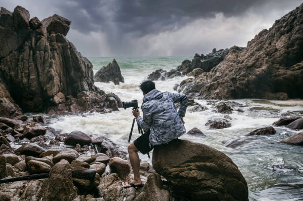 istock Photographer man holding with camera, tripod and facing the rainstorm on seashore 1348097520