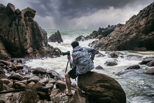 Proud middle aged photographer man holding with camera, tripod and facing the rainstorm on seashore