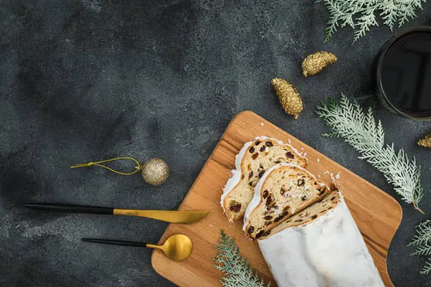 Christmas composition with stollen cake on a wooden board, coffee cup and cutlery on dark background. Flat lay