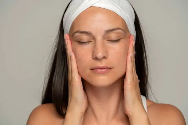 a young woman with closed eyes massaging her face on a gray background
