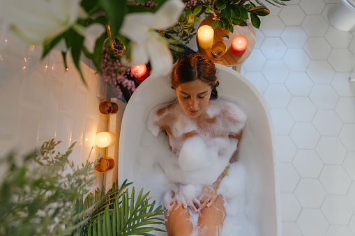 Photo of a young woman taking a bubble bath; relaxing and having a spa day at home.