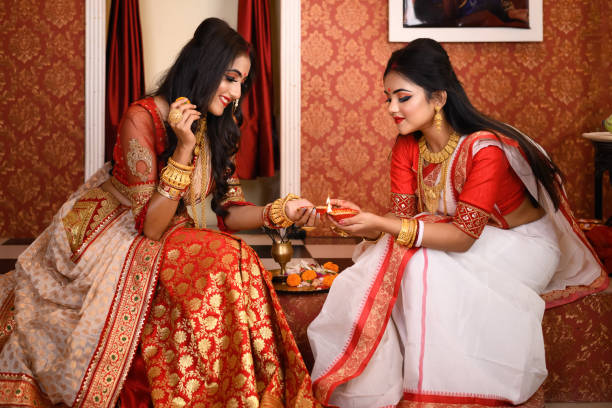Two Indian women wearing traditional saree, gold jewellery and bangles sitting at home with flower and light diyas in decorative background. Indian festival, culture, occasion, religion and fashion. stock photo