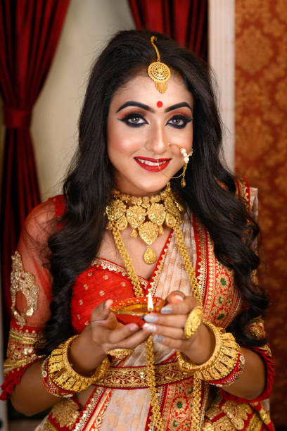 Portrait of pretty young Indian woman wearing traditional saree, gold jewellery and bangles holding diya in hands on decorative background. Indian culture, occasion, religion and fashion. stock photo