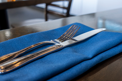 Angled view of a fork and knife placed delicately on a royal blue cloth napkin on a wooden table inside a restaurant