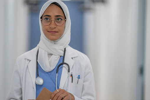 A female Muslim Doctor stops to smile casually for a portrait while walking the hallway of a hospital.  She is wearing blue scrubs, a white lab coat and Hijab and is holding a clipboard.