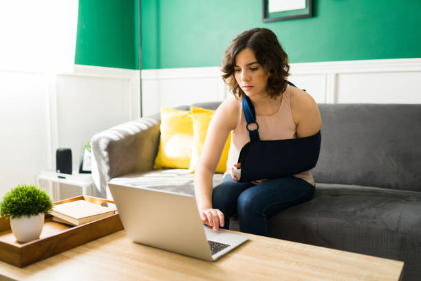 Woman with a fractured arm using the laptop Gorgeous young woman with an arm sling typing on the laptop after a having an accident and a broken bone orthopedic cast stock pictures, royalty-free photos & images