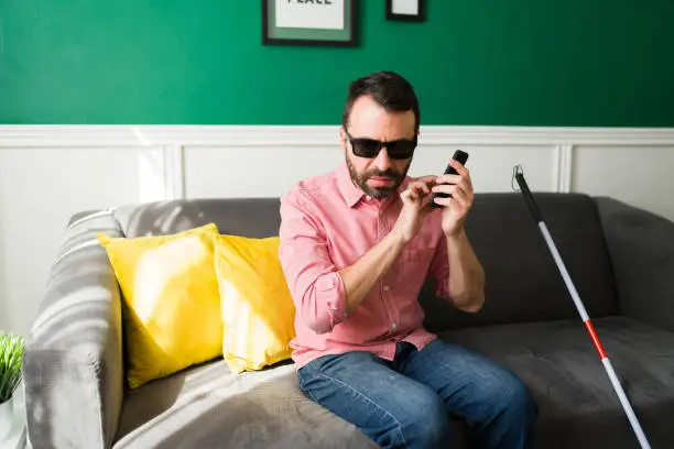 Smart blind man texting on his smartphone. Attractive man with a disability using his phone while sitting on the couch