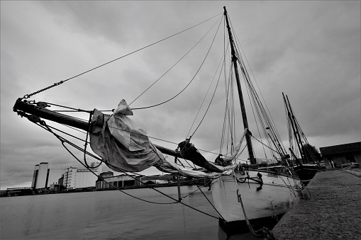 Two sailing boats are docked in the city. Black and white picture, with a cloudy sky.