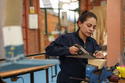 Female college student planning wood with a hand plane while building furniture in a manufacturing class - education concepts