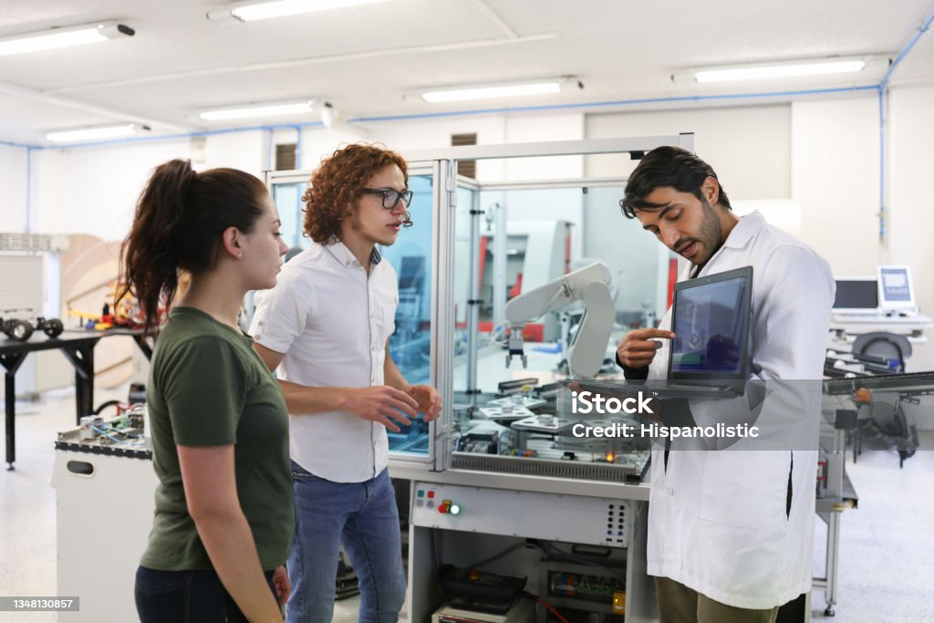Teacher explaining something to a couple of college students in a robotics class Latin American teacher explaining something to a couple of college students in a robotics class using a laptop computer - STEM education concepts Artificial Intelligence Stock Photo