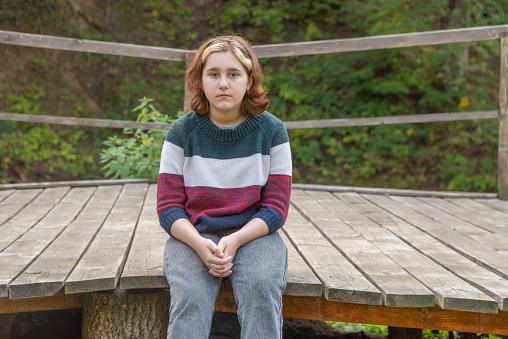 a teenage girl with a short haircut sits on a long wooden bench in nature and looks straight ahead in early autumn.