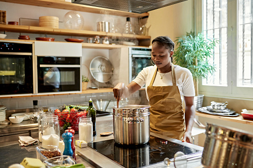 Waist-up view of woman in apron standing at kitchen workstation glass-ceramic stove and concentrating on stirring pot.