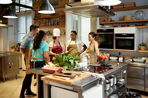 Wide angle view of male and female corporate colleagues standing around kitchen island covered with vegetables and listening to instructor in white toque.