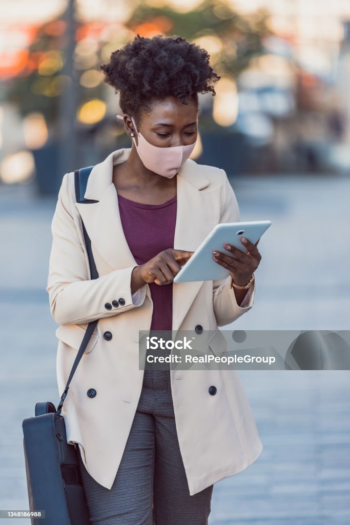 Young black woman in  a protective face mask working on the street using a tablet PC Young attractive elegant businesswoman with protective mask using touchpad while standing against modern office building. Protective Face Mask Stock Photo