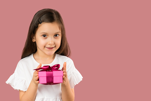 Front view of a cute little girl is holding a pink gift box in her hands with a nice smile in front of pink background.