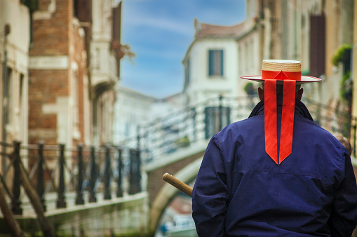 The gondolier sails on a boat along the canal, the gondolier's hat with red ribbons as a symbol of Venice. Small canal old colorful houses and bridge, Venetian lagoon, Venice, UNESCO world heritage site, Veneto, Italy, Europe.