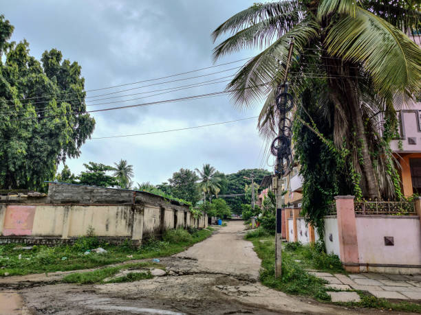 istock grey color road constructed from cement surrounded with coconut trees and Indian styled homes in cloudy climate. No people around the road. Dramatic dark clouds covered the sky. 1348198121