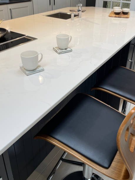 istock Image of modern kitchen design with central island breakfast bar, white composite corian countertop and coffee mugs on distressed painted square wooden coasters, padded, wood and chrome high backed bar stools 1348203494