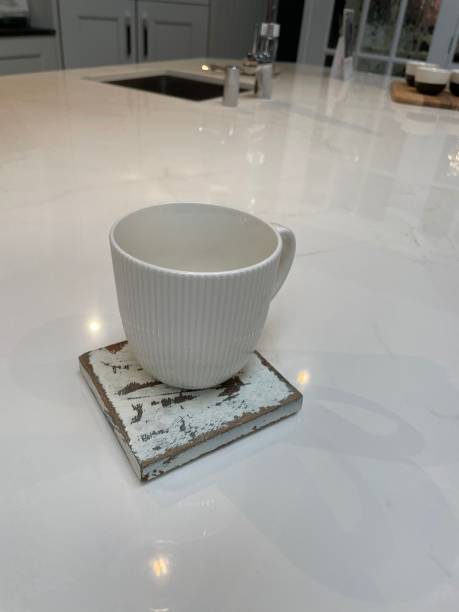 istock Close-up image of modern kitchen design with central island breakfast bar, white composite corian countertop and coffee mug on distressed painted square wooden coasters, cabinets, sink and mixer tap background 1348203552