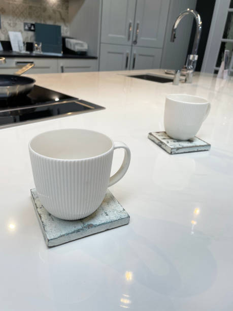 istock Close-up image of modern kitchen design with central island breakfast bar, white composite corian countertop and coffee mugs on distressed painted square wooden coasters, cabinets, sink and mixer tap background 1348203587