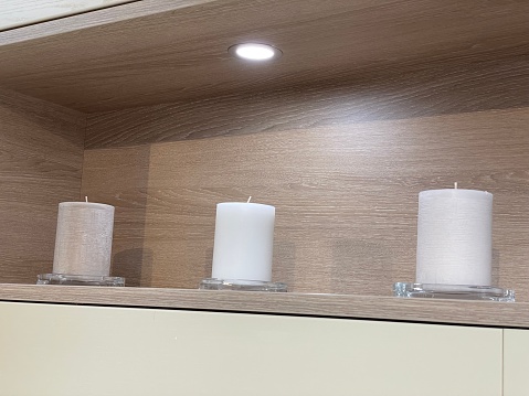 Stock photo showing close-up view of modern kitchen wall shelf. On the shelf is a row of three white pillar candles in glass candle holders.