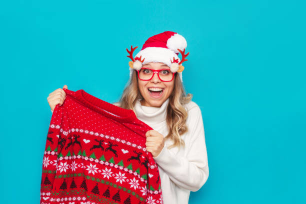 a young woman in a white warm sweater and a red santa hat holds red sweater with deer pattern. christmas and new year concept - ugly sweater imagens e fotografias de stock