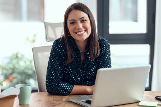 donna d'affari sorridente che lavora con il laptop mentre guarda la fotocamera nel moderno ufficio di avvio. - sorridere foto e immagini stock