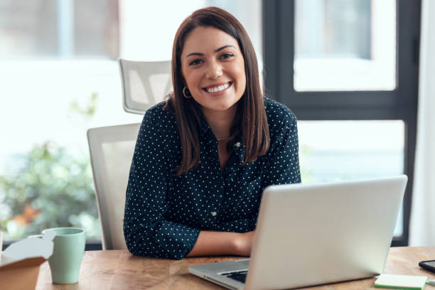 smiling business woman working with laptop while looking at camera in modern startup office. - woman stockfoto's en -beelden