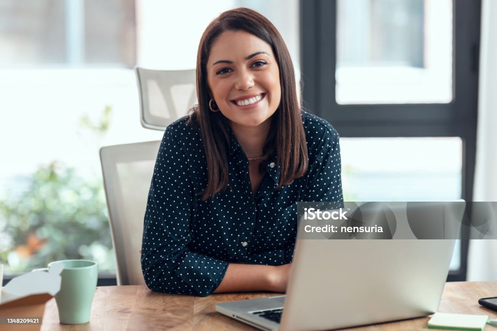 Smiling business woman working with laptop while looking at camera in modern startup office. Shot of smiling business woman working with laptop while looking at camera in modern startup office. Women Stock Photo