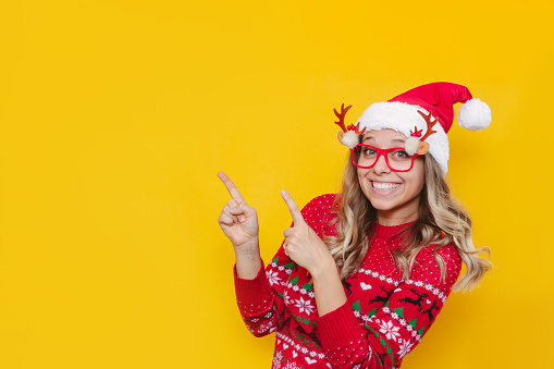 A young smiling blonde woman in a red Christmas sweater, glasses with deer antlers and a red Santa's hat points at copy space for text or design with her fingers isolated on a color yellow background