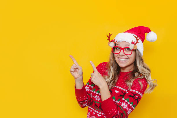 A young woman in a red deer sweater and a Santa hat points at copy space. Christmas and New Year concept A young smiling blonde woman in a red Christmas sweater, glasses with deer antlers and a red Santa's hat points at copy space for text or design with her fingers isolated on a color yellow background christmas sweater photos stock pictures, royalty-free photos & images