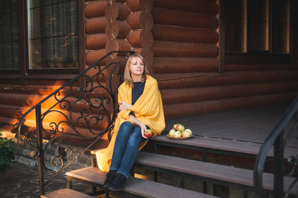 A beautiful blonde woman sits on the steps of the porch of a large wooden house. The woman is wrapped in a blanket, smiling and holding an apple in her hand. A place for text. Village eco-friendly life. Autumn relaxation. A woman over forty. Healthy lifestyle. Warm village autumn. cottagecore stock pictures, royalty-free photos & images