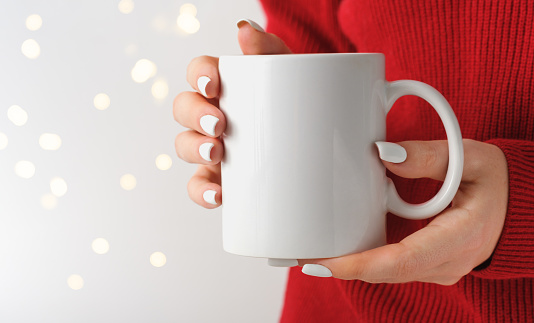 Female hands hold a mockup white tea mug in a red winter sweater, gold bokeh. For your logo and design. Christmas New Year concept. Template, empty