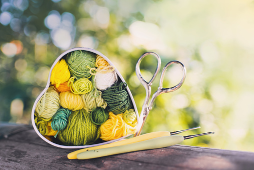 Heart-shaped box with knitted multi-colored skeins of yarn,crochet hooks , scissors on wooden background in the garden on spring day. Crochet and knitting. Women's working space.