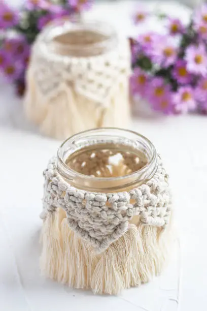 Small glass vase, jar, candleholder with macrame cover. Autumn flowers blurred on background. Boho style. Bohemian home decor. Wedding accessory.