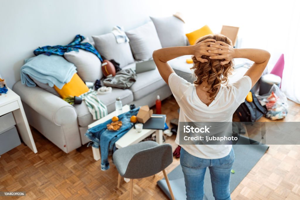 Housewife at modern home on sunny day looking at dirty room. Rear view of Mature housewife standing while preparing for cleaning at home. Messy Stock Photo
