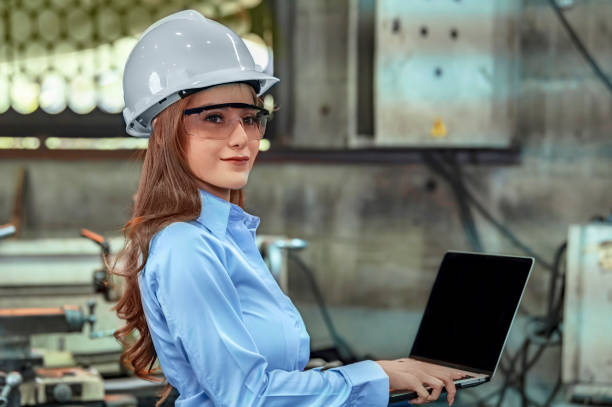 An industrial engineer wearing a white hard helmet while working on laptop. An inspector or supervisor may be present within the factory. stock photo